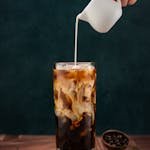 A hand pours milk into a tall glass of iced coffee next to coffee beans.