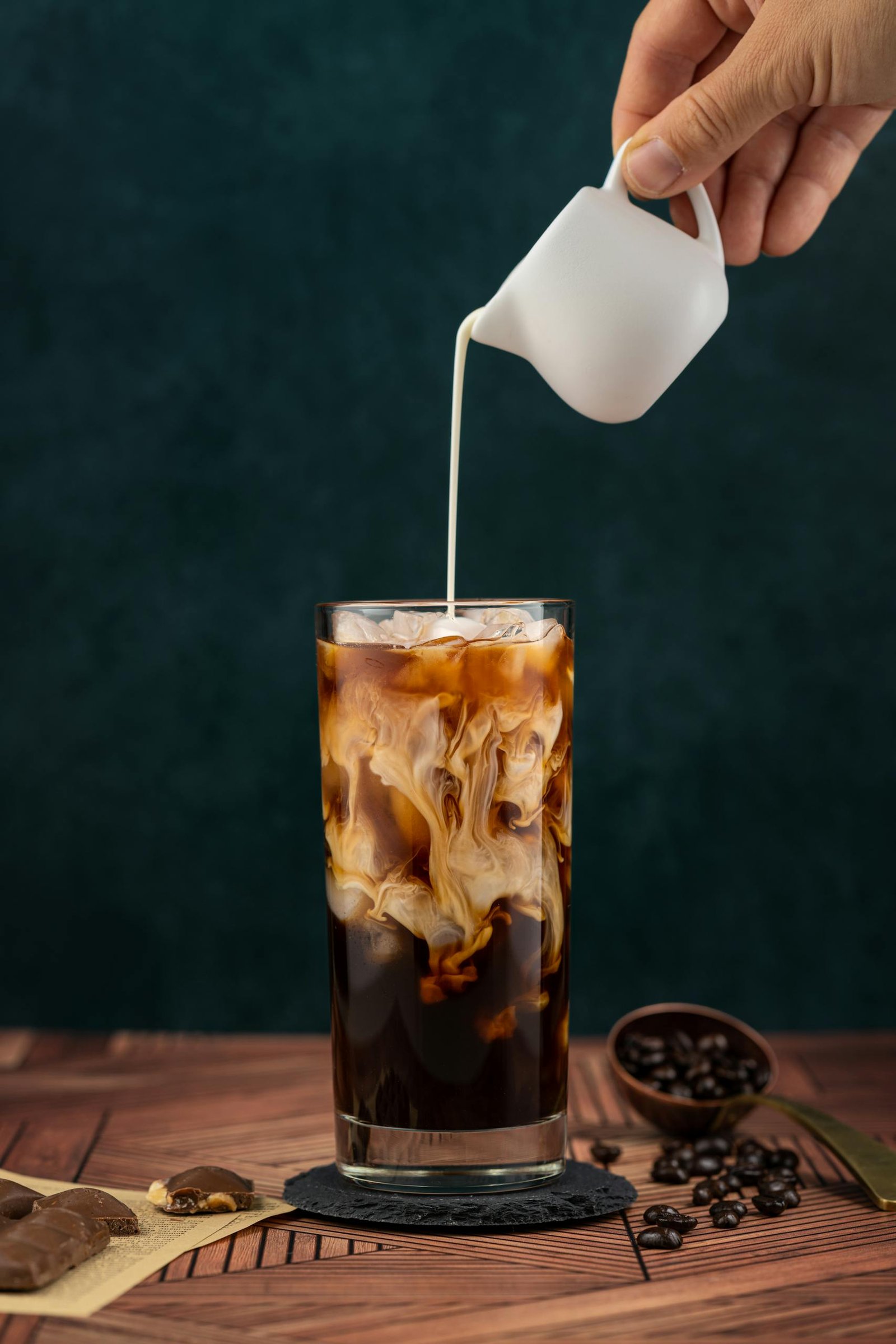 A hand pours milk into a tall glass of iced coffee next to coffee beans.