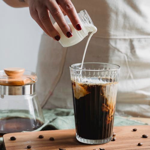 A person pours milk into a glass of iced coffee, surrounded by coffee beans, creating a captivating swirl.